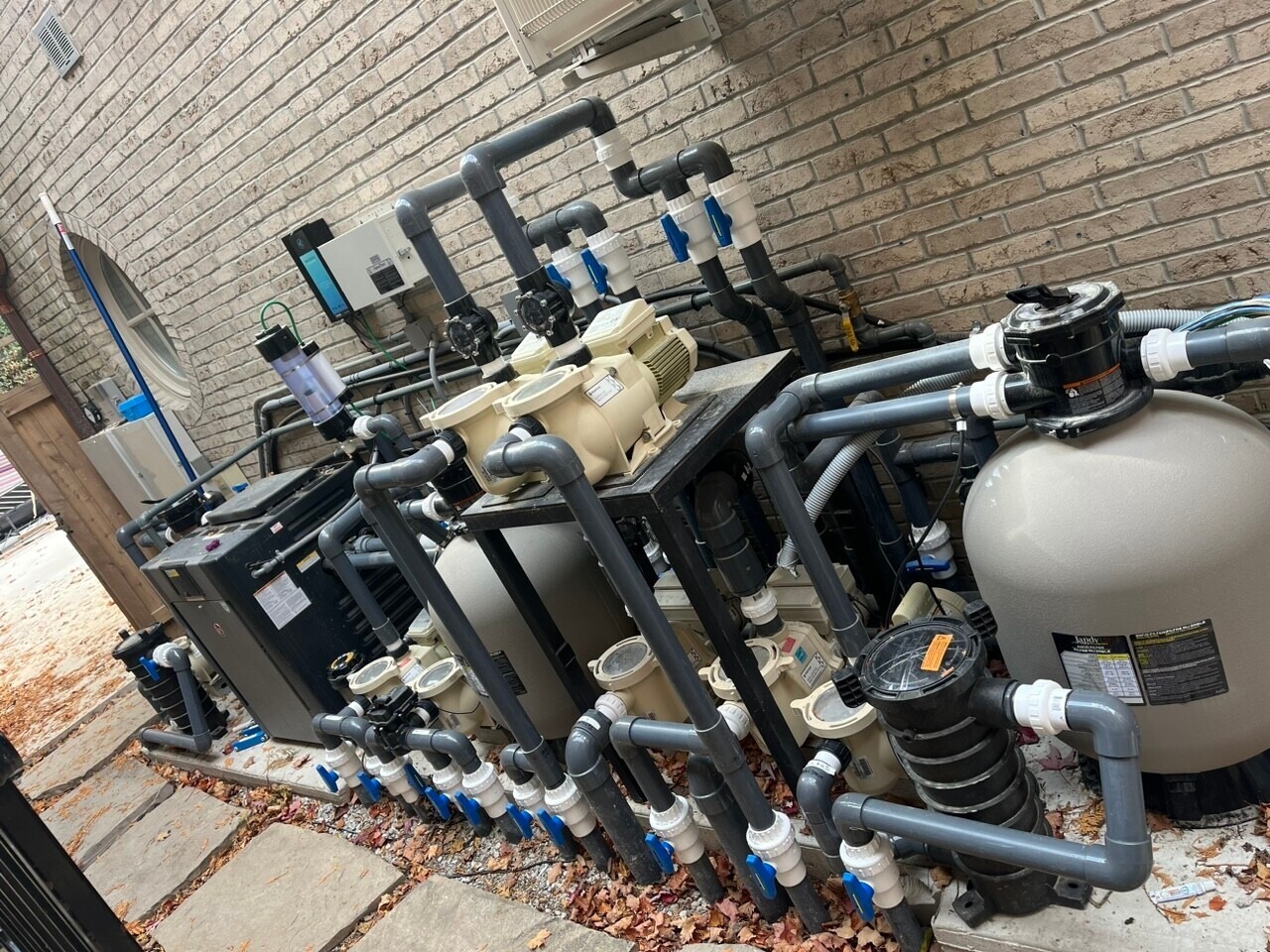 The image shows an outdoor pool mechanical system with pumps, filters, and pipes, positioned against a brick wall on a paved area.