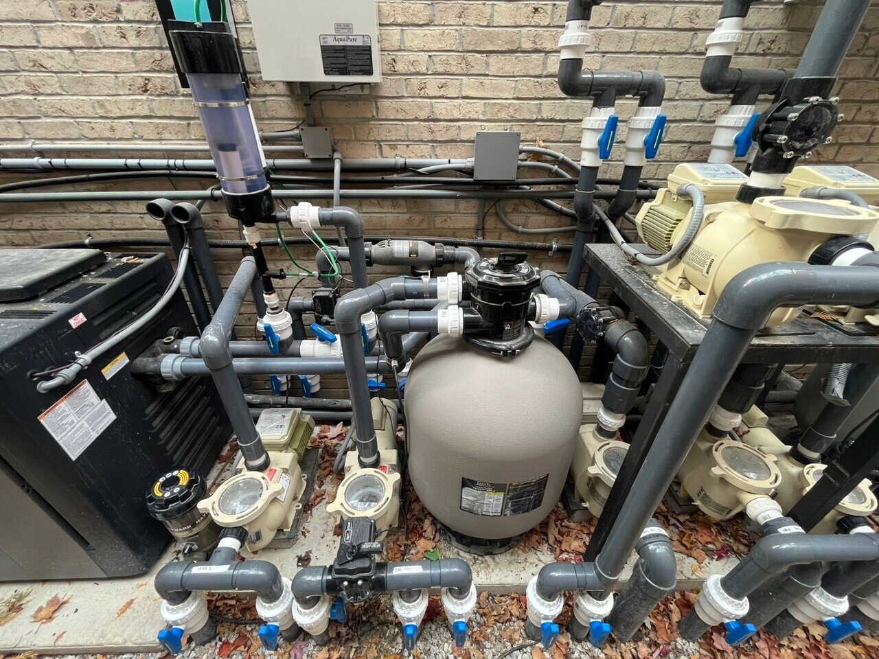 An intricate pool filtration system with pipes, pumps, and valves is set against a brick wall, appearing complex and engineered for water circulation.