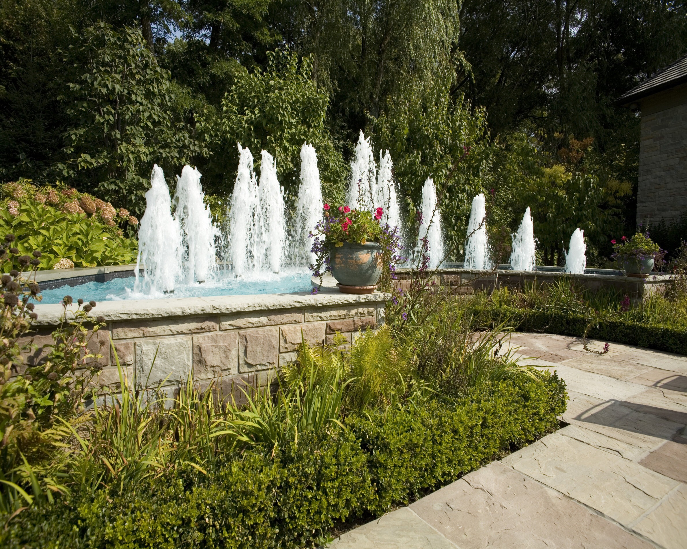 A serene garden featuring a series of water jets creating a linear fountain against lush greenery, bounded by a stone patio with flowering plants.