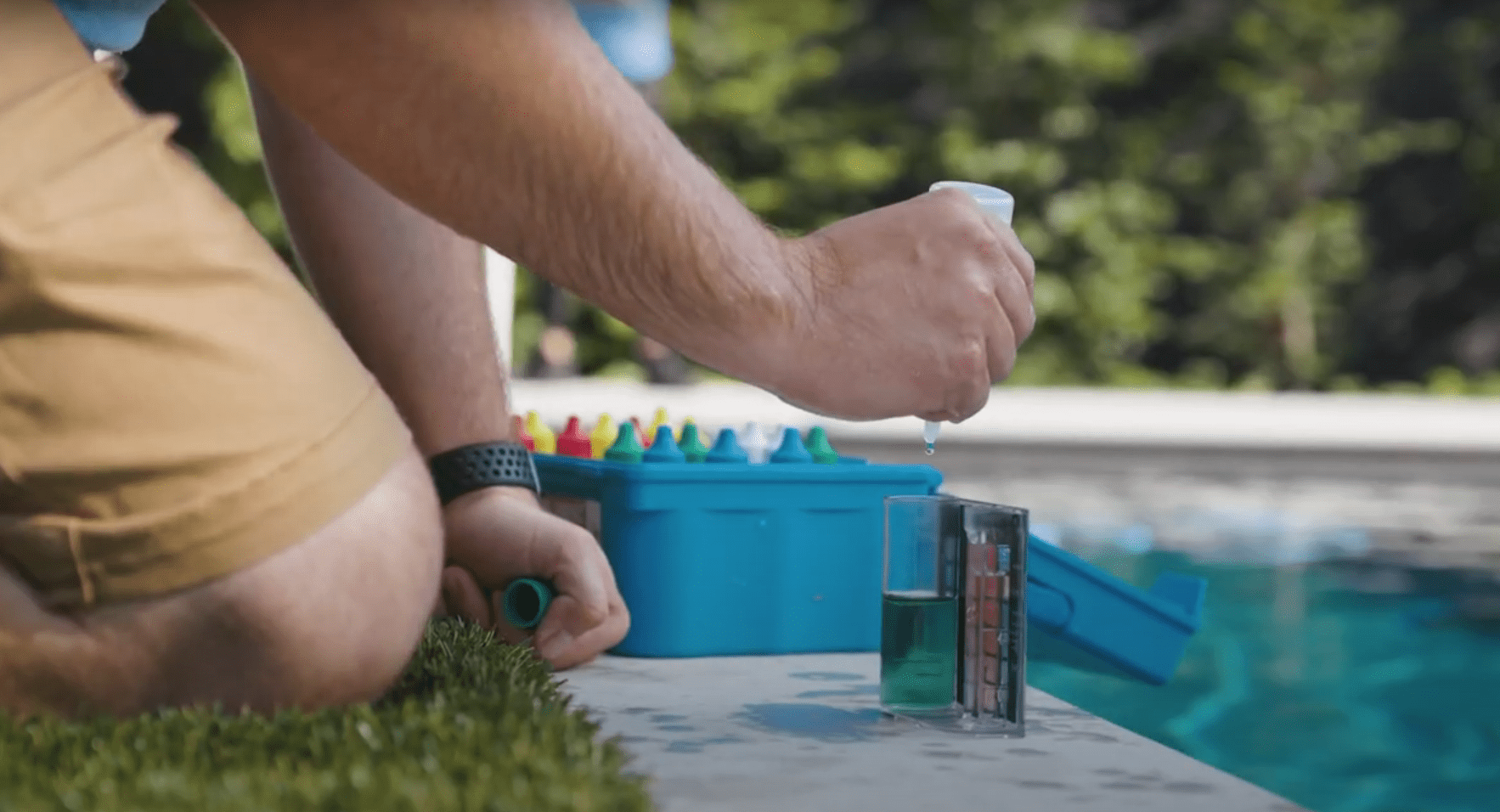 A person is testing pool water quality using a kit, dripping liquid into a measuring vial, kneeling by a swimming pool on a sunny day.