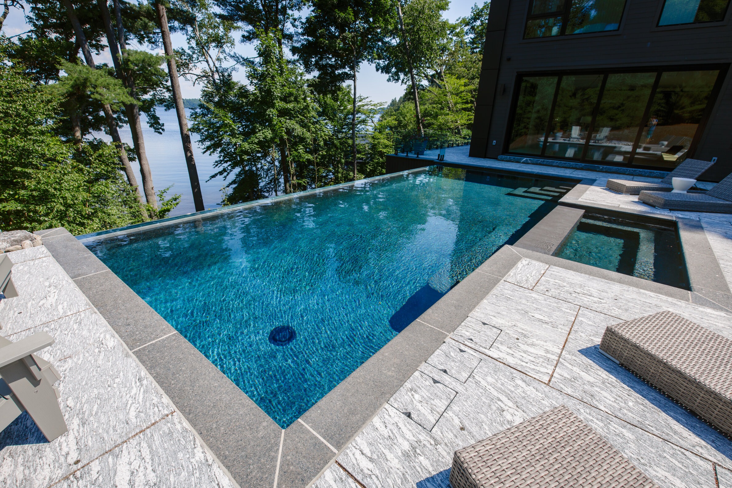 An infinity pool overlooking a scenic lake surrounded by trees, adjacent to a modern house with large glass windows and a stone patio.