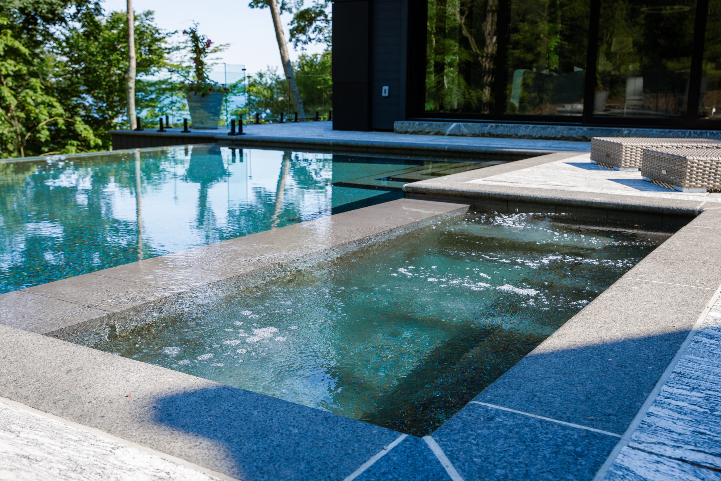 Outdoor pool and hot tub on a sunny day, surrounded by trees and lounge seating. Glass doors reflect the serene, natural environment.