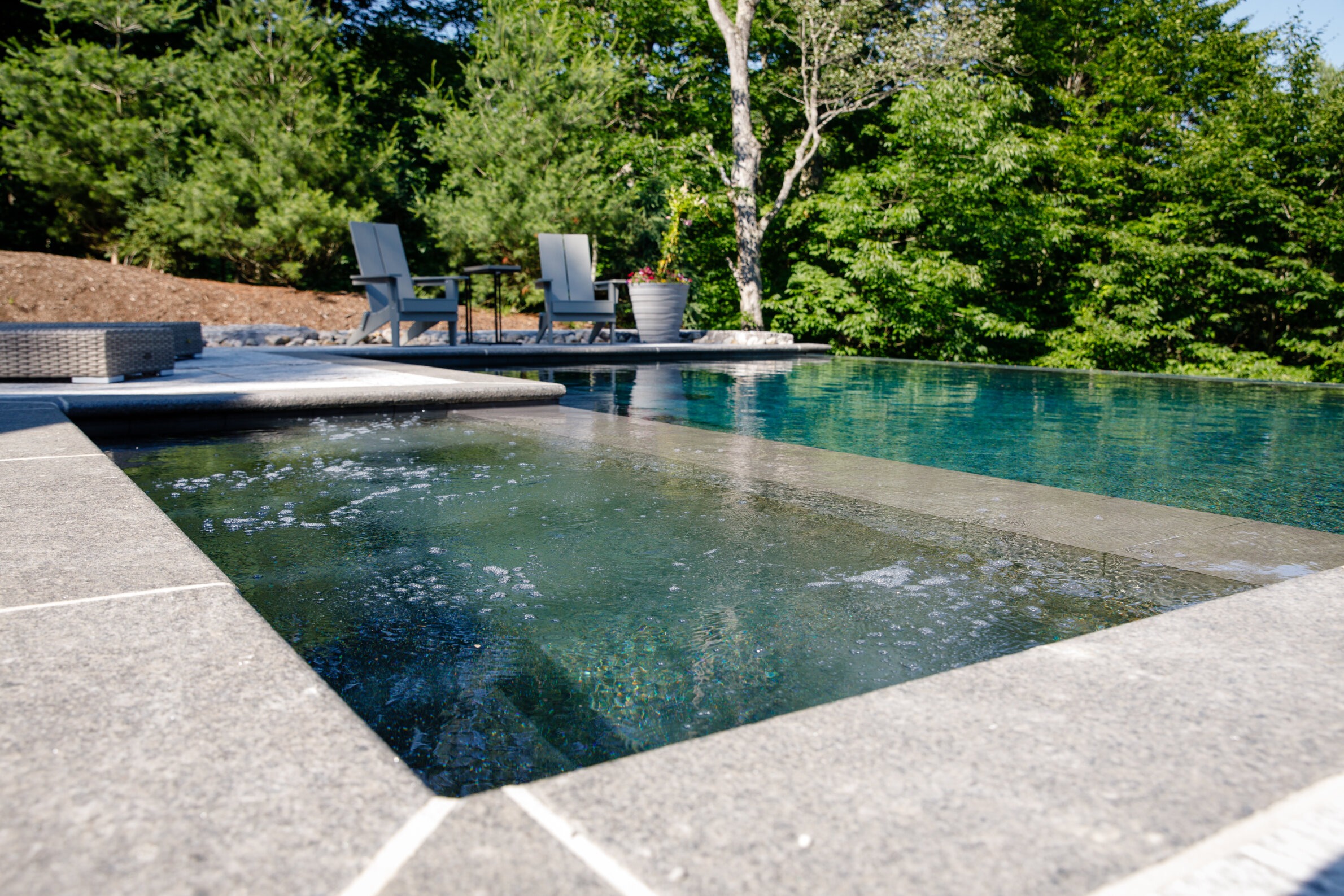 A tranquil outdoor pool with seating area, surrounded by lush greenery and trees, reflecting a peaceful, sunny atmosphere. No people are visible.