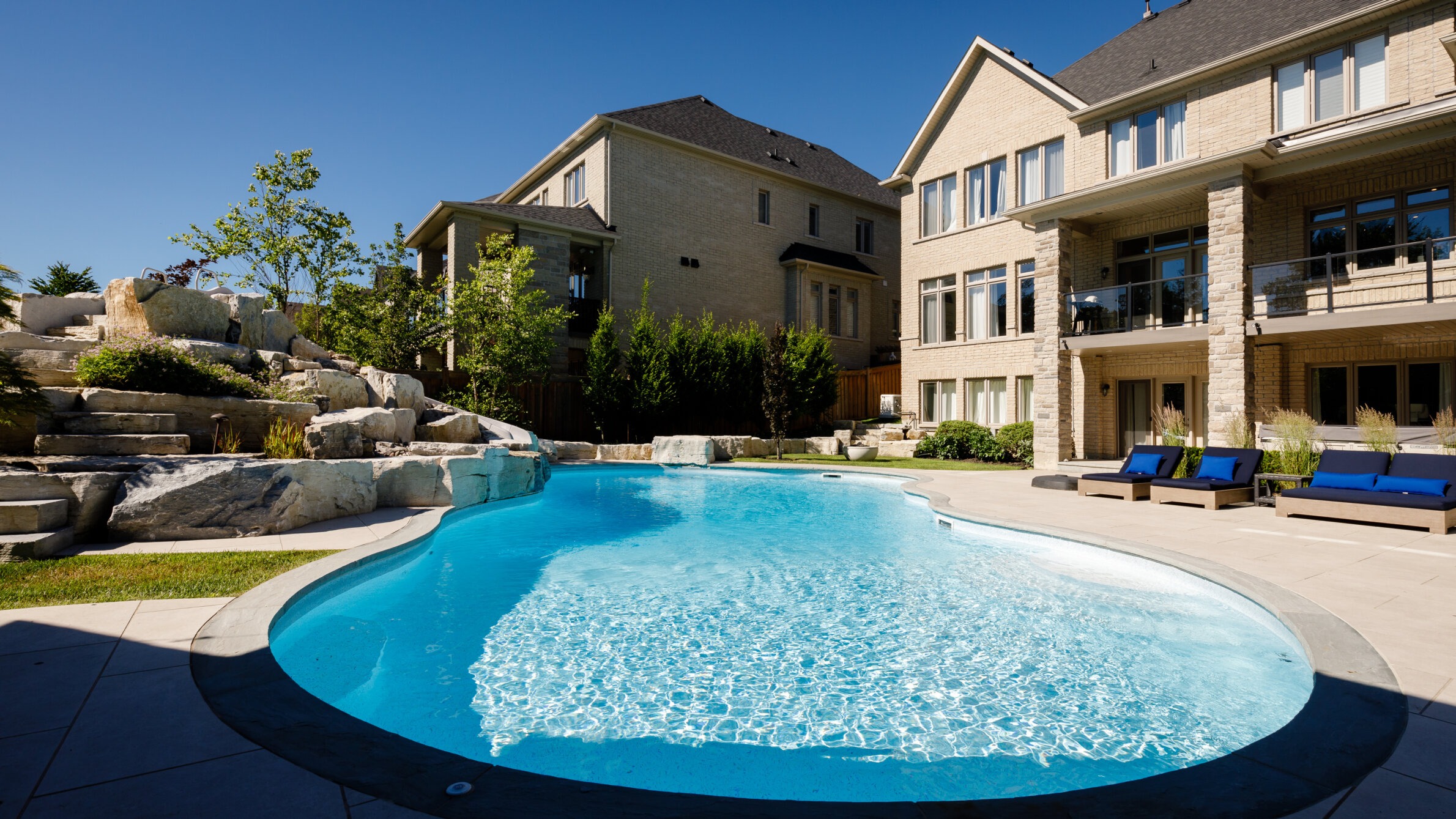 A luxurious backyard features a large swimming pool, stone landscaping, and a multi-story house with large windows under a clear blue sky.