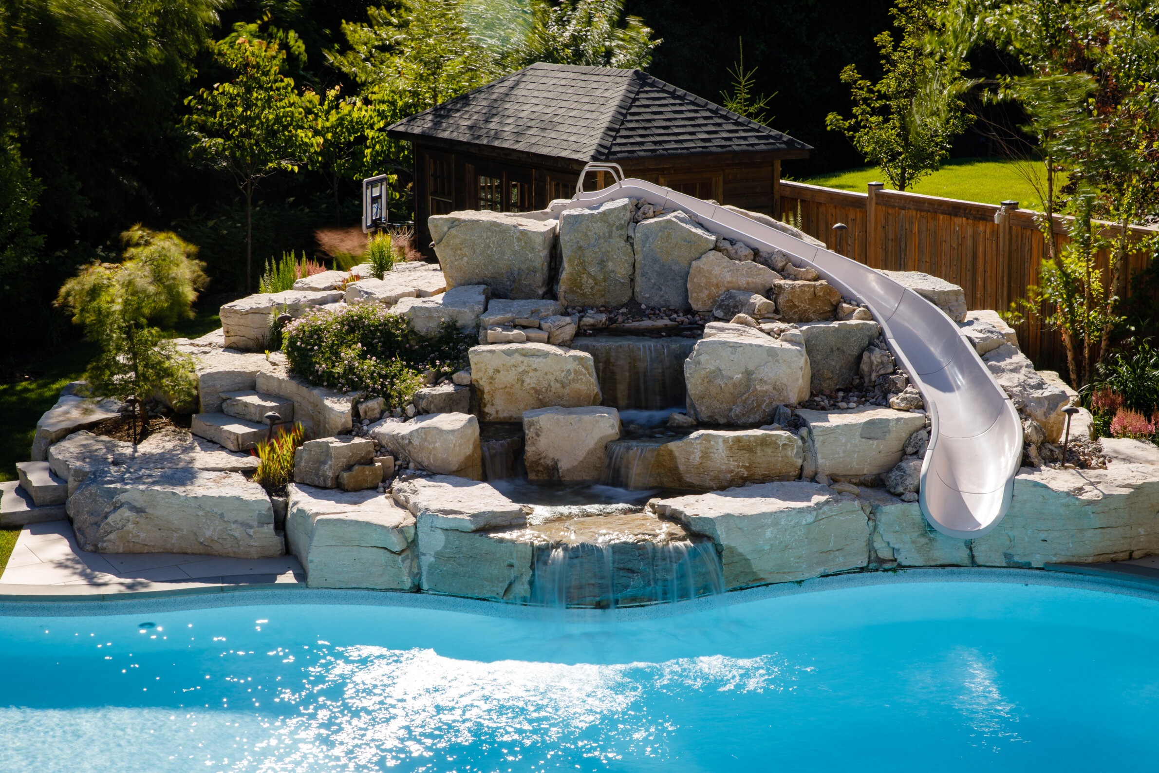 A backyard pool features a rock waterfall and slide, surrounded by lush greenery and a small wooden structure in the background.