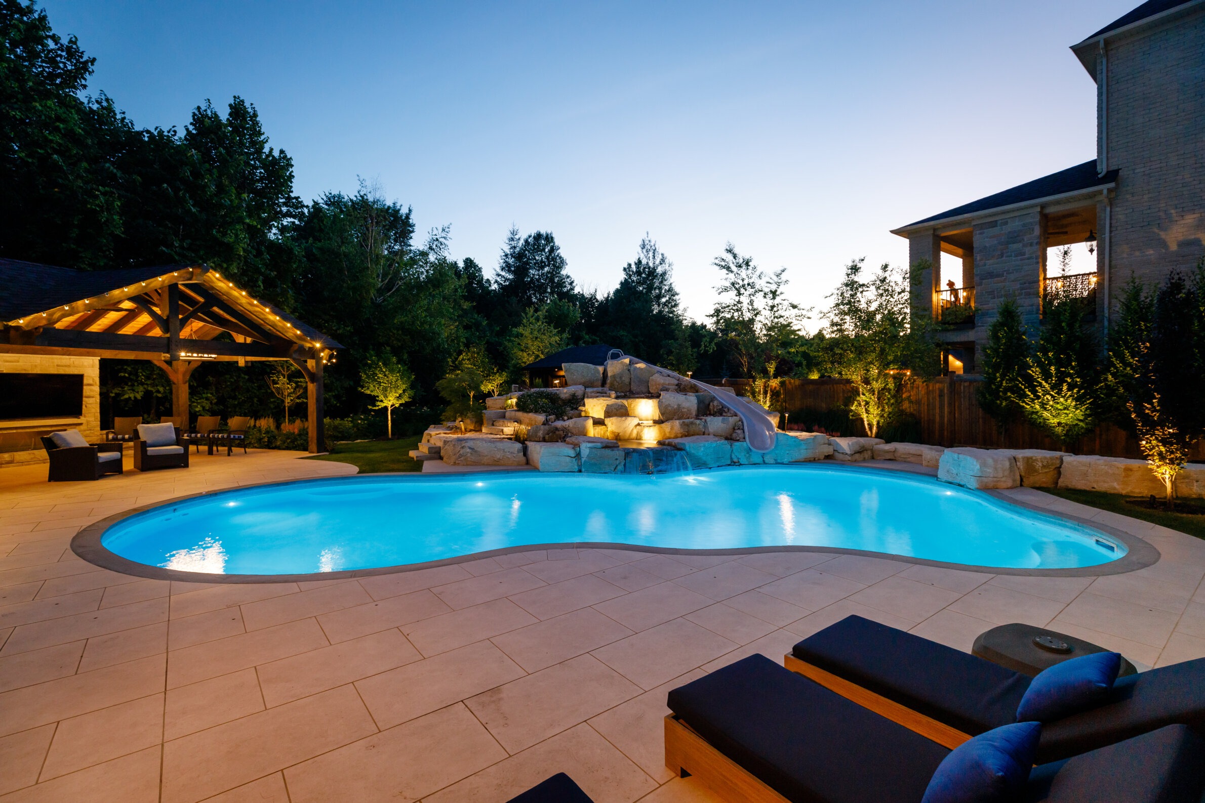 A serene backyard setting with a glowing pool, lounge chairs, wooden gazebo, and surrounding trees during twilight.