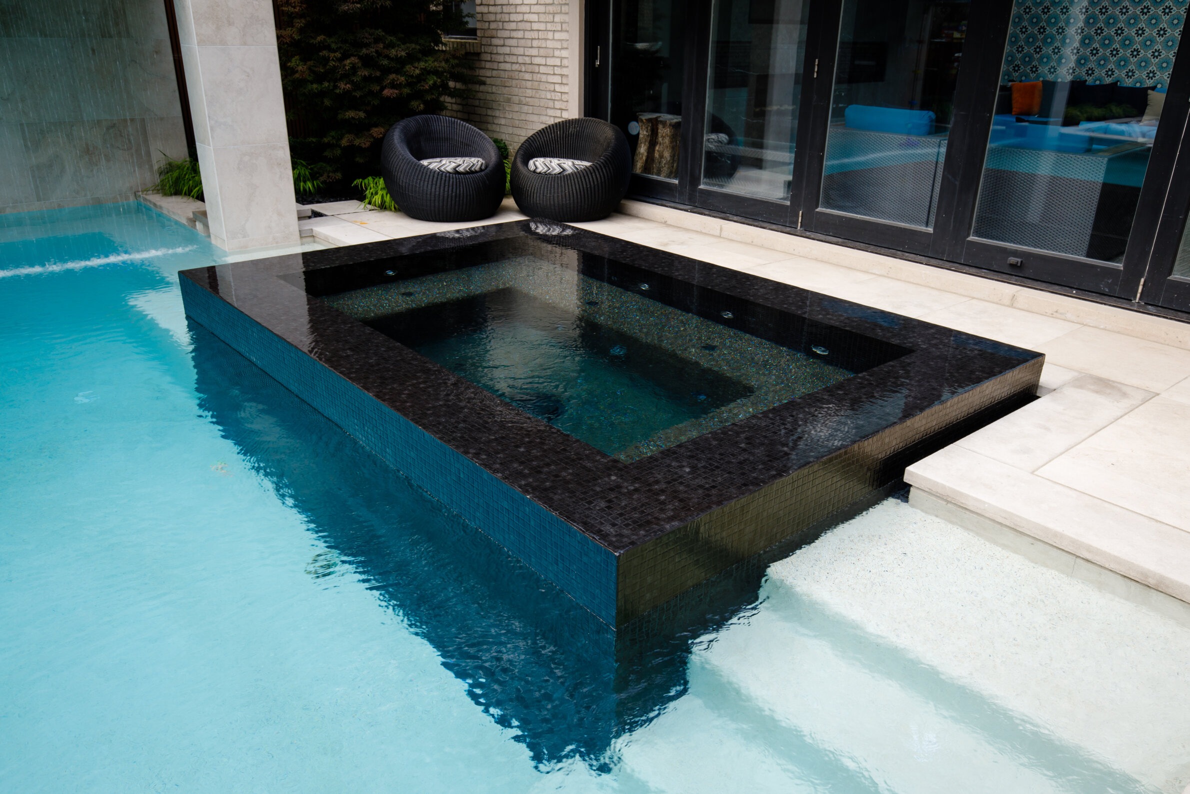 A modern outdoor pool with a black-tiled hot tub, surrounded by lounge chairs, located next to a building with large glass doors.