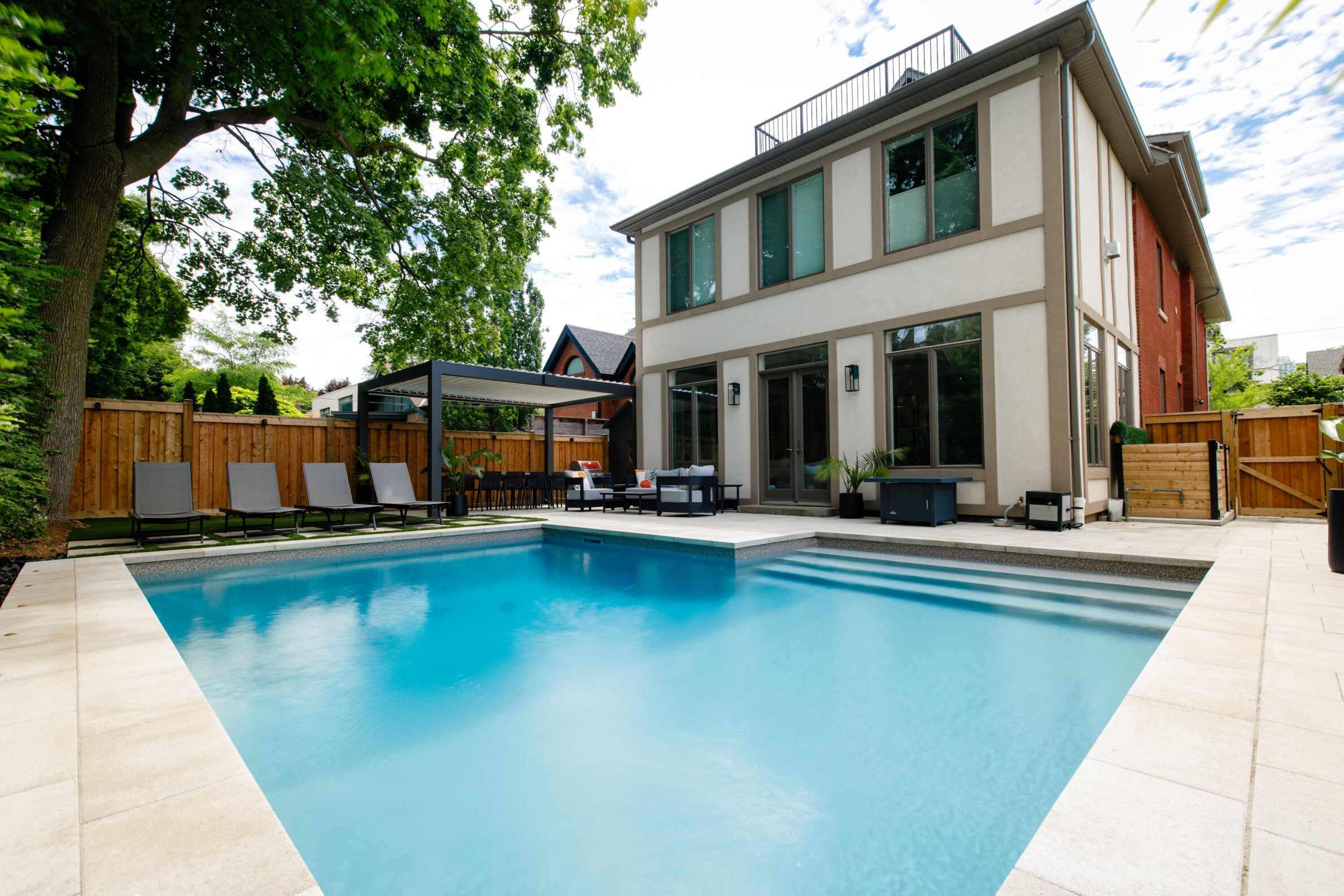 Modern house with large windows next to a backyard swimming pool, surrounded by lounge chairs and greenery, under a partly cloudy sky.