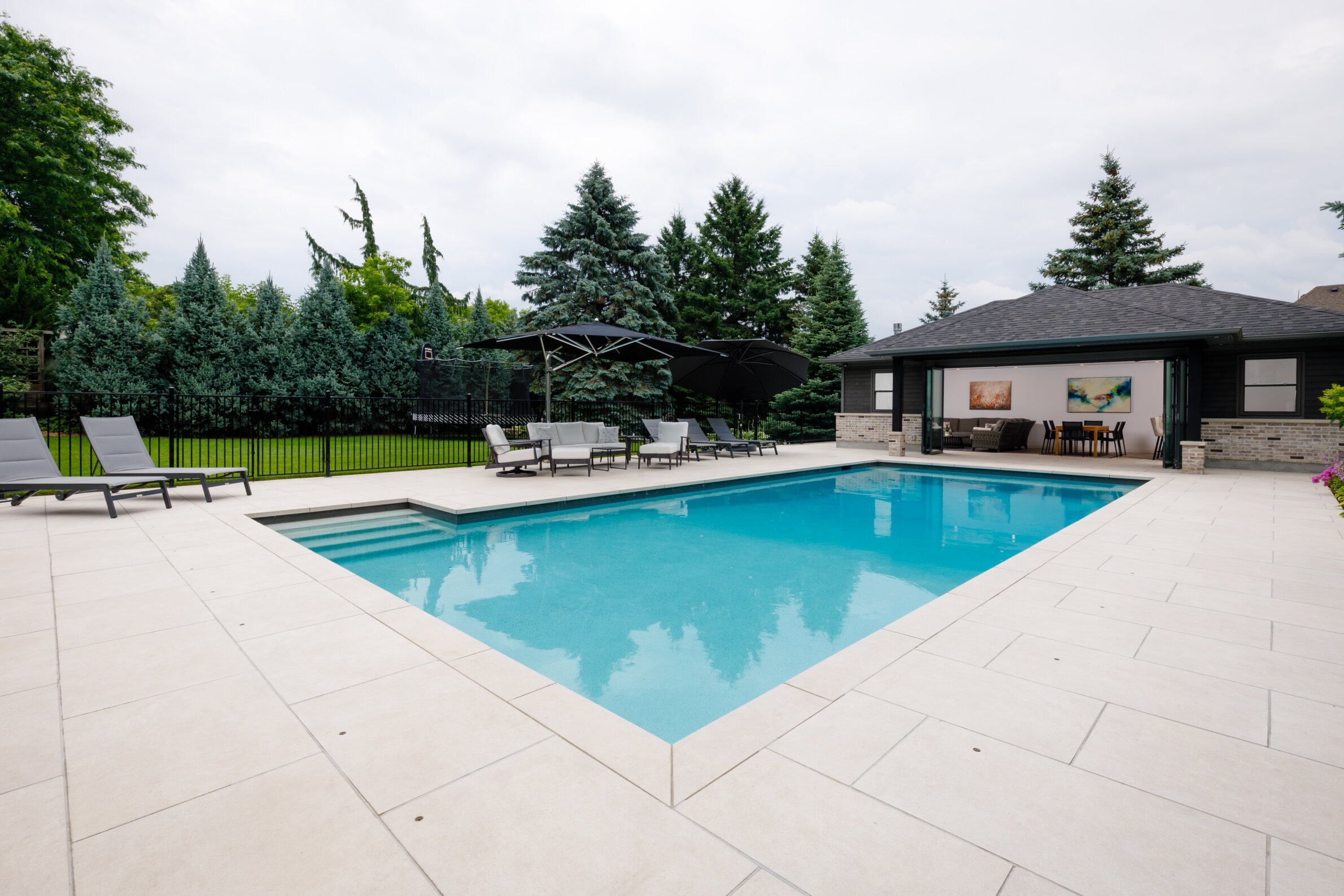 Modern backyard with a rectangular swimming pool, surrounded by lounge chairs and trees. A pool house sits nearby under a cloudy sky.