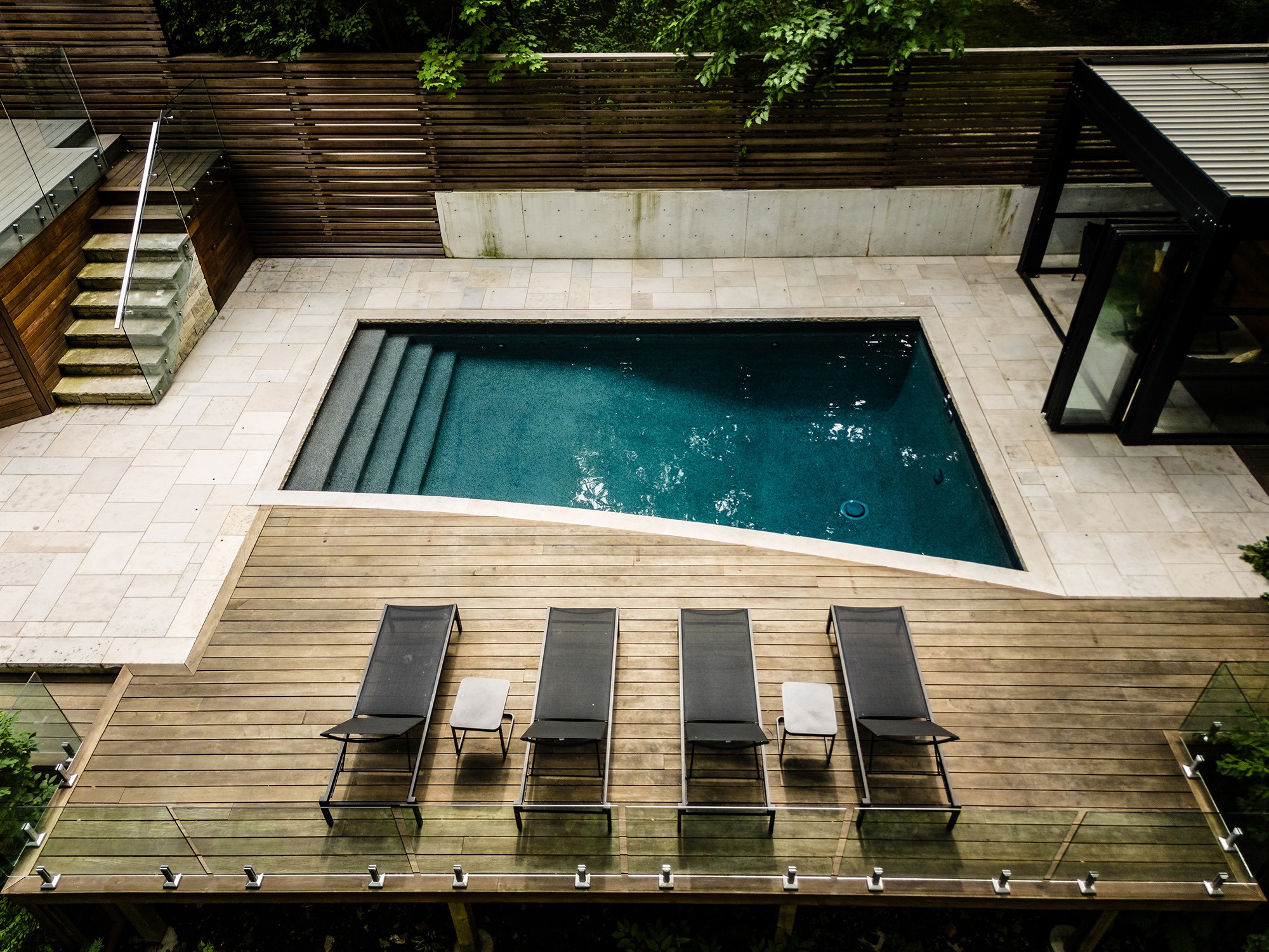 Modern backyard with rectangular pool, surrounded by wood and stone decking. Four lounge chairs line the deck. No people or landmarks visible.