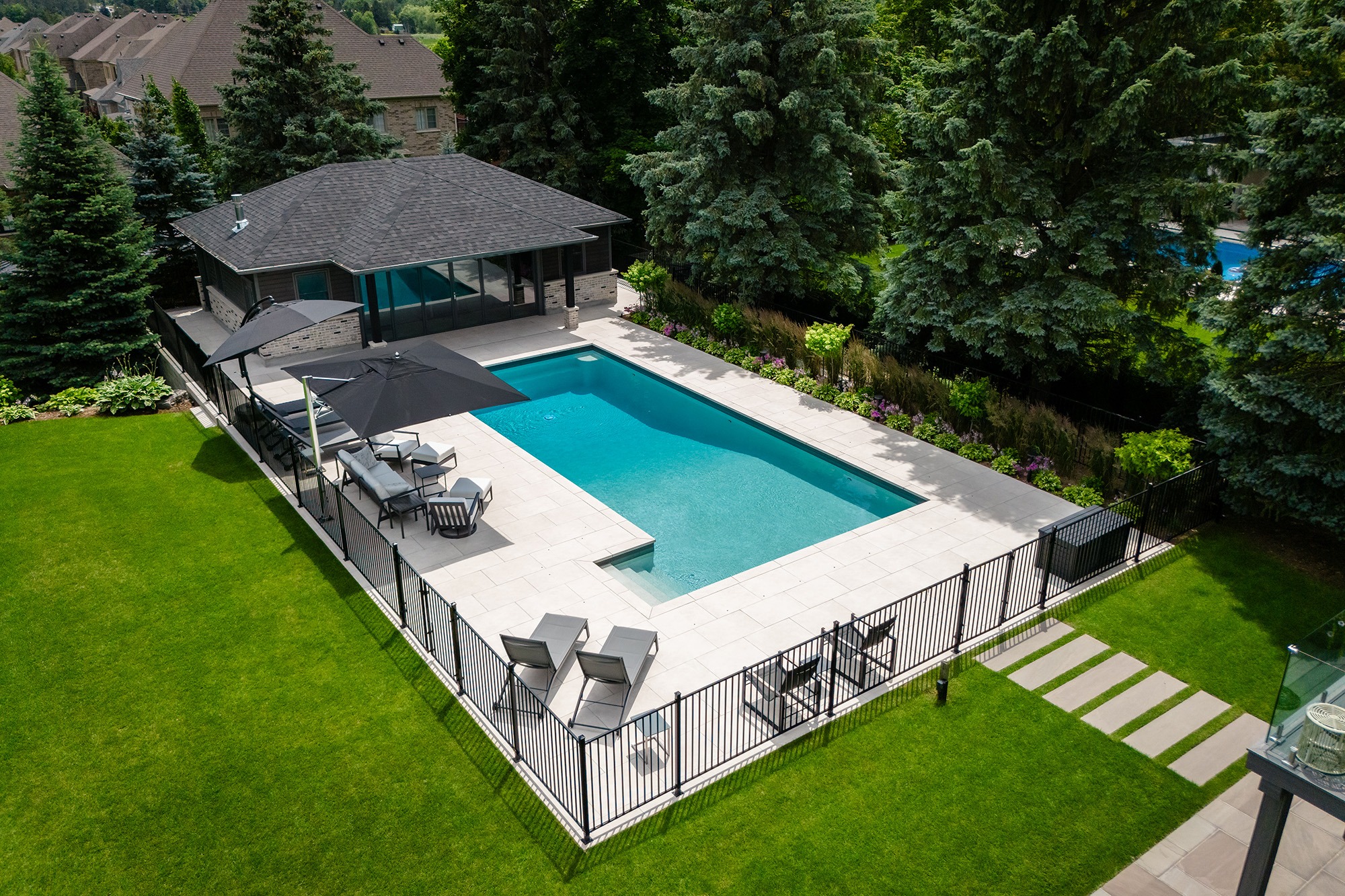 Aerial view of a private backyard with a modern pool, patio furniture, green lawn, surrounded by trees and a wooden fence.