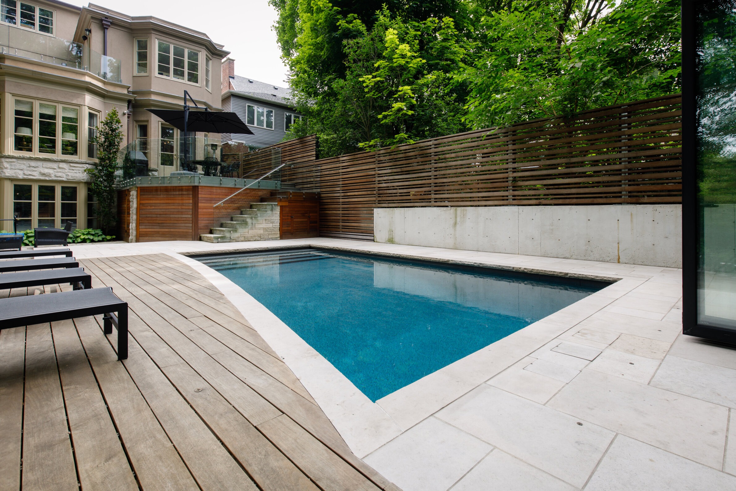 A modern backyard pool is surrounded by wooden and stone decking, adjacent to a contemporary house with large windows and lush greenery.